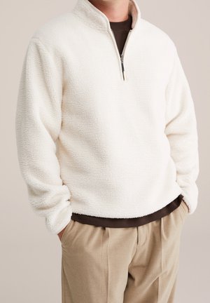 Mężczyzna ubrany w biały polar z zamkiem na ćwiartkę, brązową koszulę pod spodem oraz beżowe spodnie, ręce w kieszeniach, neutralne tło.