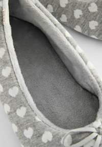 Grijze slipper met een wit hartpatroon, zachte stoffen voering en een platte, geveerde binnenkant. Kenmerkt zich door een klein strikdetail aan de voorkant.