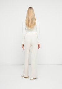 Biała krótka bluzka z długimi rękawami o prążkowanej teksturze, w połączeniu z jasnobeżowymi spodniami typu wide-leg. Białe sneakersy z brązową podeszwą są widoczne.