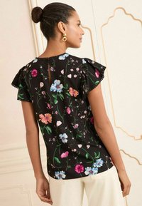 Blusa nera a fiori con maniche corte a balze. Presenta fiori colorati in tonalità di rosa, blu e verde, con un dettaglio a oblò sul retro.