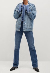 Osoba v ošuntělé světle modré džínsové bundě, modrém pleteném svetr, tmavě modrých džínách a černých botách s úzkou špičkou.