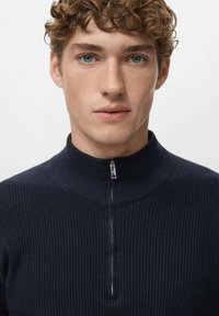 Marineblauer Rippstrickpullover mit hohem Kragen und Halbreißverschluss. Der Stoff wirkt weich mit einer strukturierten Passform, ohne sichtbare Muster oder Verzierungen.