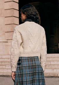 Chunky cream cable-knit sweater with a cropped design, paired with a blue and brown plaid skirt featuring side buttons. Textured surfaces are visible.