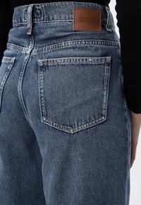Gros plan sur la poche arrière d'un jean en denim bleu avec une étiquette en cuir marron de la marque Drykorn sur la ceinture, porté avec un haut noir.