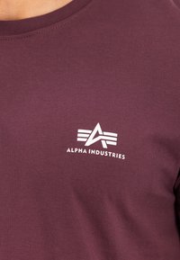 Bordeauxrotes Hemd mit Alpha Industries-Logo in Weiß auf der Brust, mit einem stilisierten "A" mit drei horizontalen Linien.