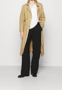 Beżowy trencz na białej koszuli, w połączeniu z czarnymi spodniami z szerokimi nogawkami i czarnymi botkami. Gładki materiał, podwójne zapięcie, minimalne detale.