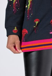 Sudadera negra con estampados de medusas coloridas, un dobladillo acanalado rosa con rayas naranjas y moradas, y una textura suave.