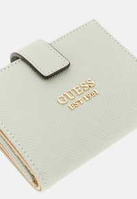 Zeleni teksturirani novčanik s kopčom na pritisak, s zlatnim logotipom "GUESS EST 1981" i kontrastnim tamnim obrubom. Kompaktni dizajn s vidljivim šavovima.