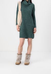 Robe tricotée verte avec un col montant en forme de capuche, des manches longues et des fentes sur les côtés. Bottes beige doublées de fourrure avec une surface texturée et des semelles robustes.
