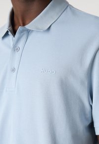 Homme portant un polo bleu clair avec un col, deux boutons et un logo "HUGO" embossé sur la poitrine gauche.