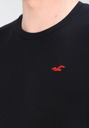 Zwarte katoenen T-shirt met ronde hals, met een klein rood geborduurd logo op de linkerborst. Gladde textuur en een minimalistisch ontwerp.