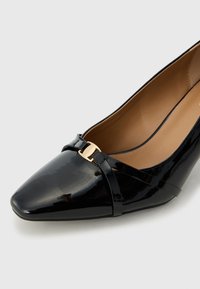 Sorte lakkpumps med spiss tå, som har en gullfarget spenne og kryssede stropper. Glatt tekstur og slank design.