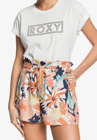 T-shirt blanche avec le logo "ROXY" au-dessus de shorts floraux colorés. Les shorts ont une taille nouée, sont en tissu léger et présentent des motifs multicolores vibrants.