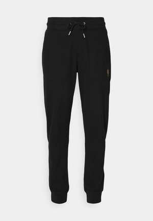 Svart joggebukse laget av myk stoff med elastisk midje og snøring, sidelommer, og en gulllogo på venstre ben.