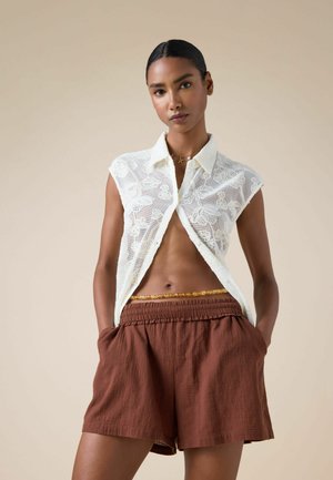 Frau trägt durchsichtiges weißes, geblümtes ärmelloses Hemd, braune Shorts mit hoher Taille und eine perlenbestückte Taillenkette, steht mit den Händen in den Taschen.
