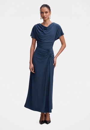 Femme portant une longue robe bleu marine froncée à manches courtes, associée à des talons noirs pointus et des boucles d’oreilles pendantes, debout devant un fond blanc.