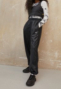 Svart vadderad crop-väst över en långärmad t-shirt med tie-dye, kombinerat med svarta mönstrade joggers och svarta atletiska skor, stående mot en texturerad vägg.