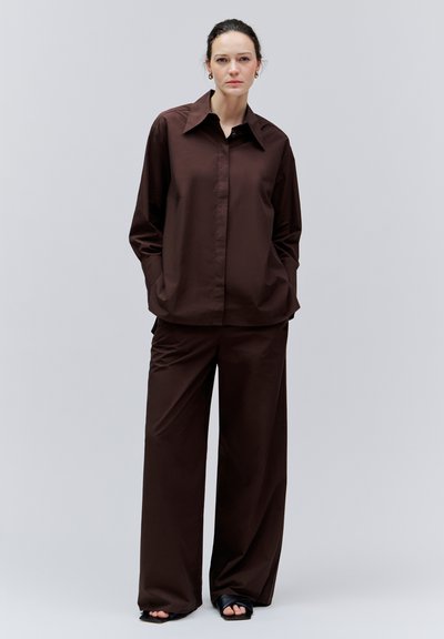 Femme debout, mains dans les poches, portant une chemise ample marron foncé et un pantalon large, avec des sandales noires à bout ouvert sur fond gris clair.