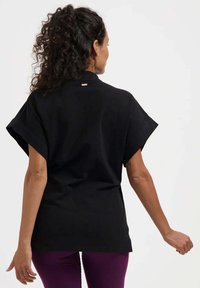 Schwarzes Kurzarmoberteil mit Kragen, das auf der Rückseite einen dezenten goldenen Akzent aufweist. Der Stoff wirkt glatt, mit einem entspannten Schnitt und einfachem Design.