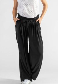 Schwarze, weit geschnittene Hose mit einem gerafften Bund und Bindegürtel vorne. Hergestellt aus weichem Stoff, kombiniert mit einem weißen Lochspitzenoberteil und weißen Sneakers.