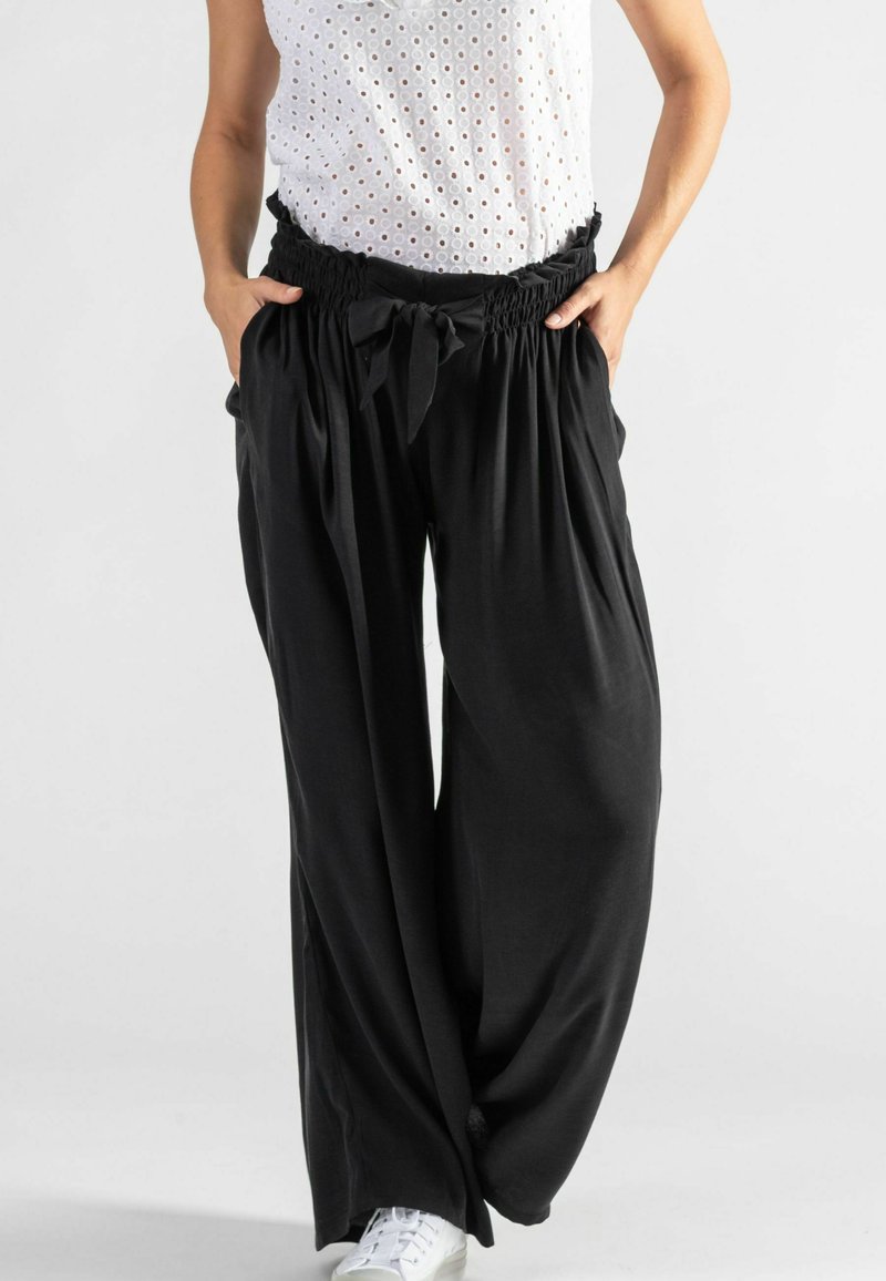 Schwarze, weit geschnittene Hose mit einem gerafften Bund und Bindegürtel vorne. Hergestellt aus weichem Stoff, kombiniert mit einem weißen Lochspitzenoberteil und weißen Sneakers.