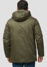 Grön huva jacka med en slät textur, med dragkedjestängning och en något längre bakstycke, bärs över blå jeans.