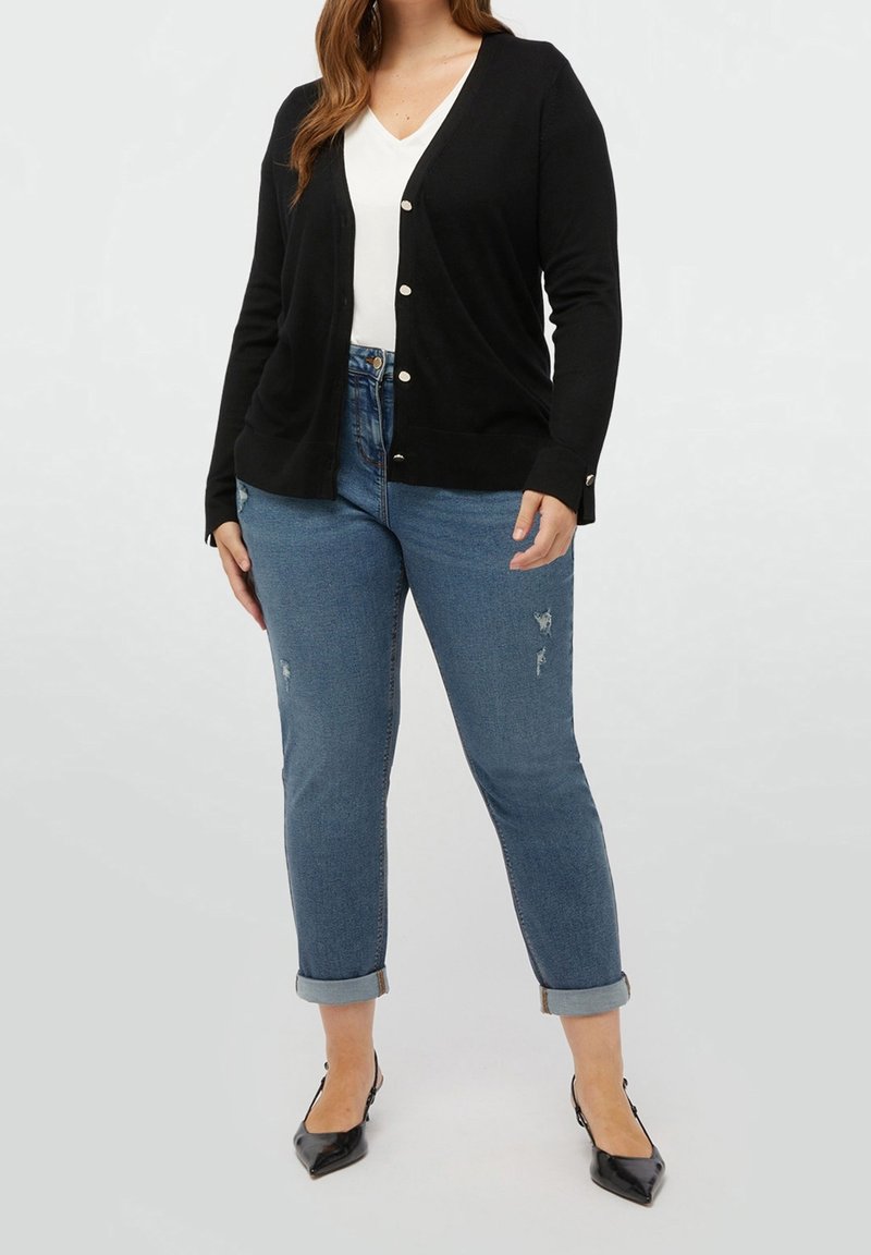 Černý kardigan s výstřihem do V, dlouhými rukávy a zapínáním na knoflíky vpředu, ve spojení s světle modrými džíny s ohrnutými lemy a poškozenými detaily.