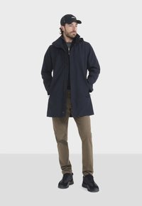 Marineblauer wasserabweisender langer Mantel mit Reißverschluss, Kapuze und sichtbaren Nähten, kombiniert mit beigen Hosen und schwarzen Sportschuhen.