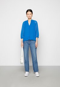 Blusa azul, semi-translúcida, com decote em V, fecho de botão à frente e mangas a três quartos. Combinada com jeans claros de perna reta e sapatilhas brancas.