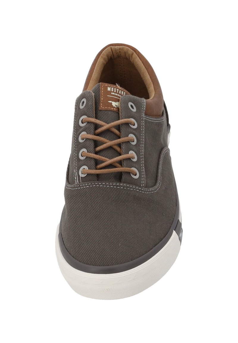 Zapatilla de lona gris con detalles en cuero marrón, que presenta cordones beige, punta redondeada y suela de goma blanca. Costuras direccionales visibles.