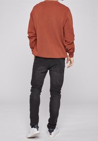 Sweter w kolorze rdzy z długimi rękawami, w parze z czarnymi dżinsami slim fit. Osoba ma na sobie białe sneakersy z ciemnymi akcentami.