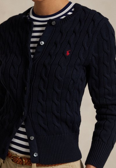 Marineblå kabelstrikket cardigan med knappestenging, med en liten rød logo på venstre bryst, båret over en stripete skjorte.