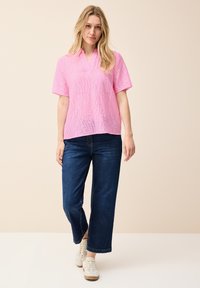 Camicetta rosa a maniche corte con scollatura a V e motivi a onda, abbinata a jeans larghi blu scuro e sneakers bianche. Design e texture minimalisti.
