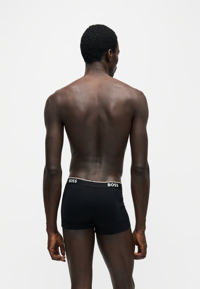 Homme debout de dos, portant un boxer noir avec une ceinture blanche BOSS sur fond blanc uni.