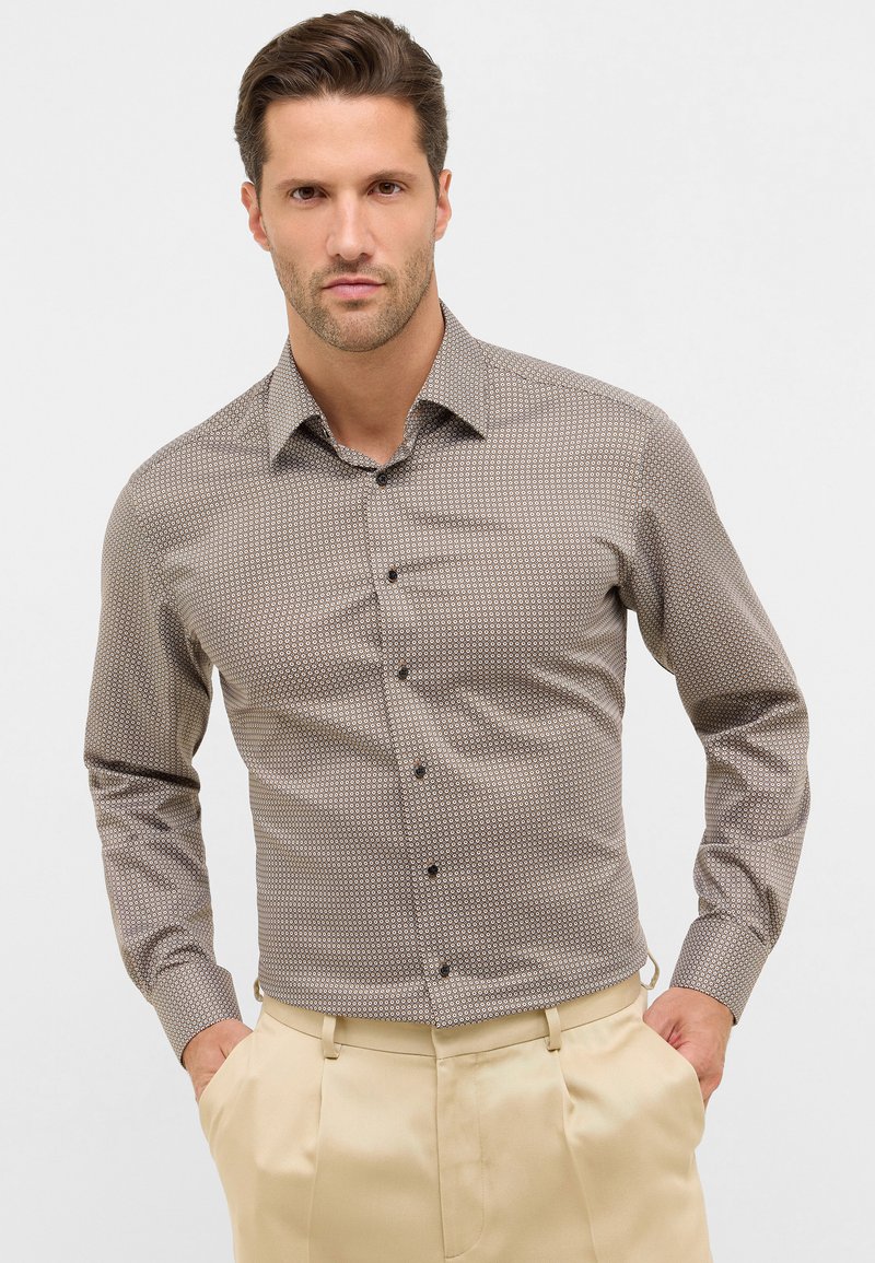 Man met kort haar, gekleed in een gestreepte langemouwen overhemden en beige broek, met de handen in de zakken, tegen een neutrale achtergrond.