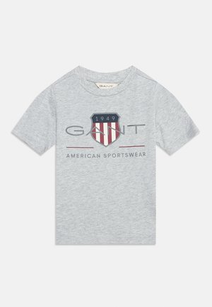 Tricou gri deschis cu mâneci scurte, cu un logo în formă de scut roșu, alb și albastru cu inscripția "1949," și textul "GANT American Sportswear" pe partea din față.