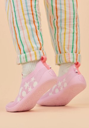Kinderfüße, die rosa Slip-on-Schuhe mit Sternenmuster und weiße Socken tragen, auf den Zehenspitzen stehen, dazu bunte gestreifte Hosen, an den Knöcheln hochgekrempelt.