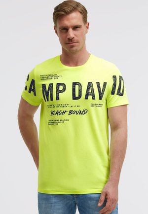 Mann trägt ein neon-gelbes T-Shirt mit dem Aufdruck "CAMP DAVID" und beach-inspiriertem Design sowie blaue Jeans, steht vor einem schlichten hellen Hintergrund.