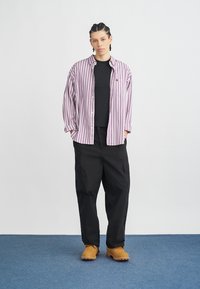 Pruhovaná fialovo-bílá košile na knoflíky přes černé tričko, doplněná volnými černými kalhotami s kapsami a hnědými dřeváky, stojící na modrém koberci.