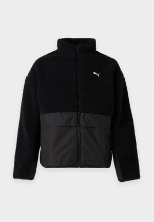 Ung mand iført en sort Puma fleecejakke, brede lysegrå jeans og sorte sneakers, stående mod en ensfarvet hvid baggrund.
