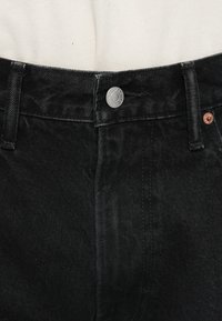 Sorte denimbukser som har metallknaplukning, bæltestropper og kontrastfarvet kobberstikning langs lommerne og sømmen.