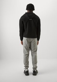 Svart hettegenser med teksturerte sidelapper, kombinert med lysegrå avsmalnende bukser og svarte joggesko, sett fra baksiden.