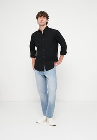 Camisa preta de botões com mangas arregaçadas; jeans azul-claro de perna reta; ténis brancos. Tecido liso e design minimalista, sem padrões.