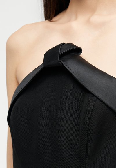 Close-up of a person wearing a black asymmetrical top with a wide, folded strap across one shoulder and a bare opposite shoulder.