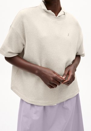 Person trägt einen hellbeigen, übergroßen Kurzarm-Sweatshirt über einem weißen Hemd mit Kragen und einem hellvioletten Rock, Hände verschränkt.