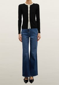Black knit cardigan with cream trim and buttons, paired with high-waisted, flared blue jeans and pointed black heels.