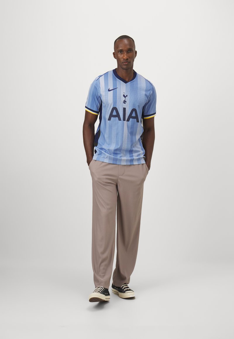 Jasnoniebieska koszulka piłkarska z pionowymi paskami, czarnym logo "AIA" oraz czarną emblematyką Spurs; zestawiona z beżowymi spodniami i czarnymi sneakerami.