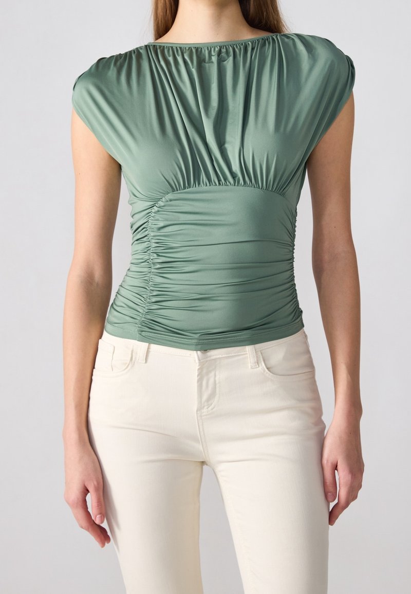 Grön, figursydd topp med rynkad tyg vid axlar och midja. Mjuk, slät textur med lätt lyster. Matchad med ljusa jeans.