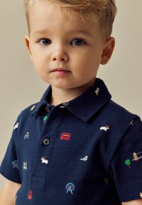Marine polo skjorte med farverige broderede motiver, herunder biler, en bus, dyr og træer. Krave-design med knaplukning.
