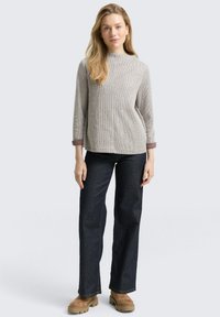 Vrouw met lang blond haar, gekleed in een lichtgrijze trui, donkerblauwe wijde jeans en tan kleurige chunky laarzen, staande tegen een effen achtergrond.